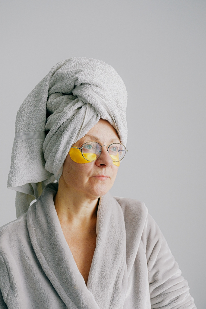 An older woman wearing undereye masks and a towel around her head.