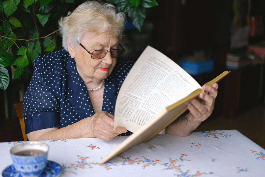 An elderly woman reading at her kitchen table.